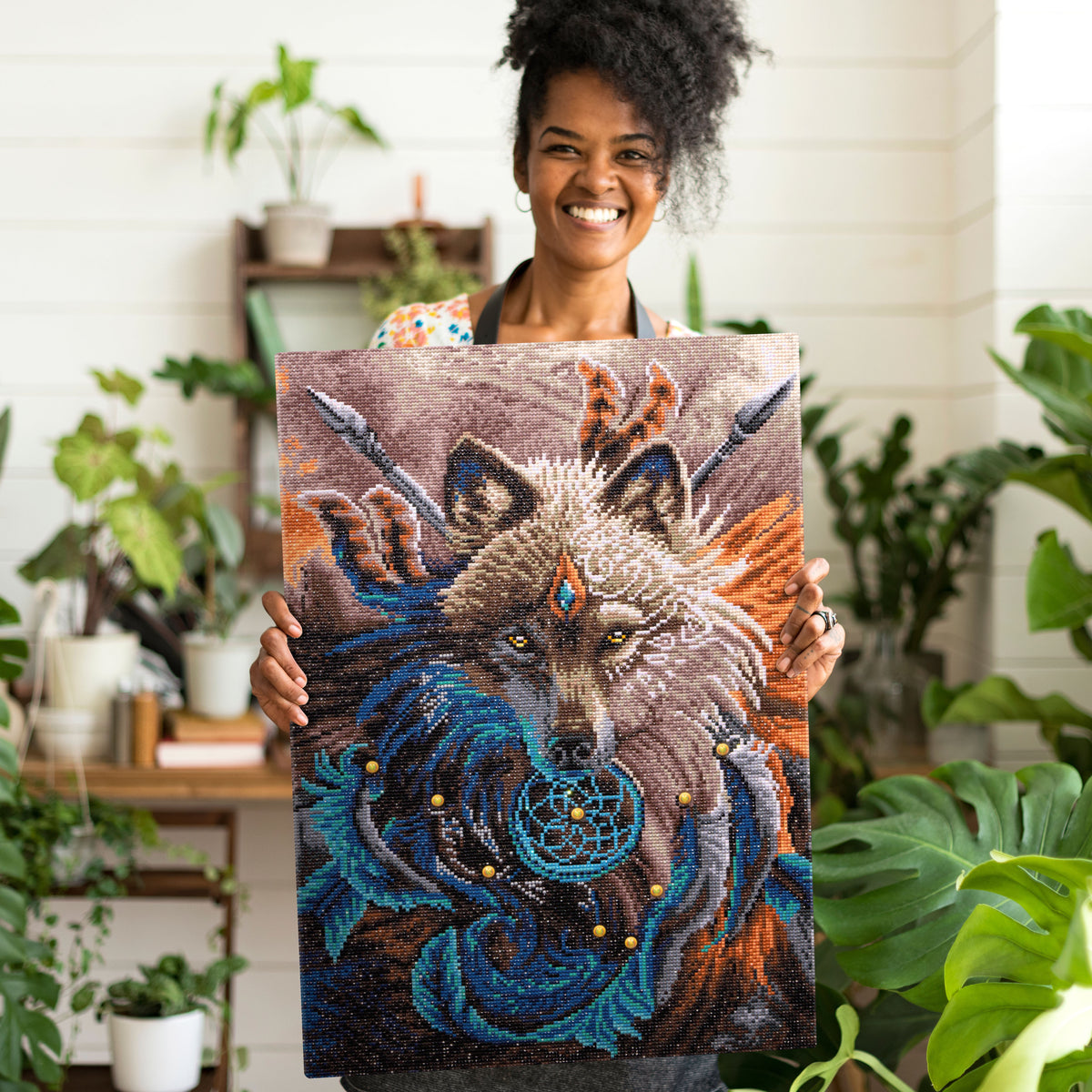 Un amateur de diamond painting souriant tenant une toile terminée de "l'attrape-rêves loup" dans une pièce lumineuse remplie de plantes.